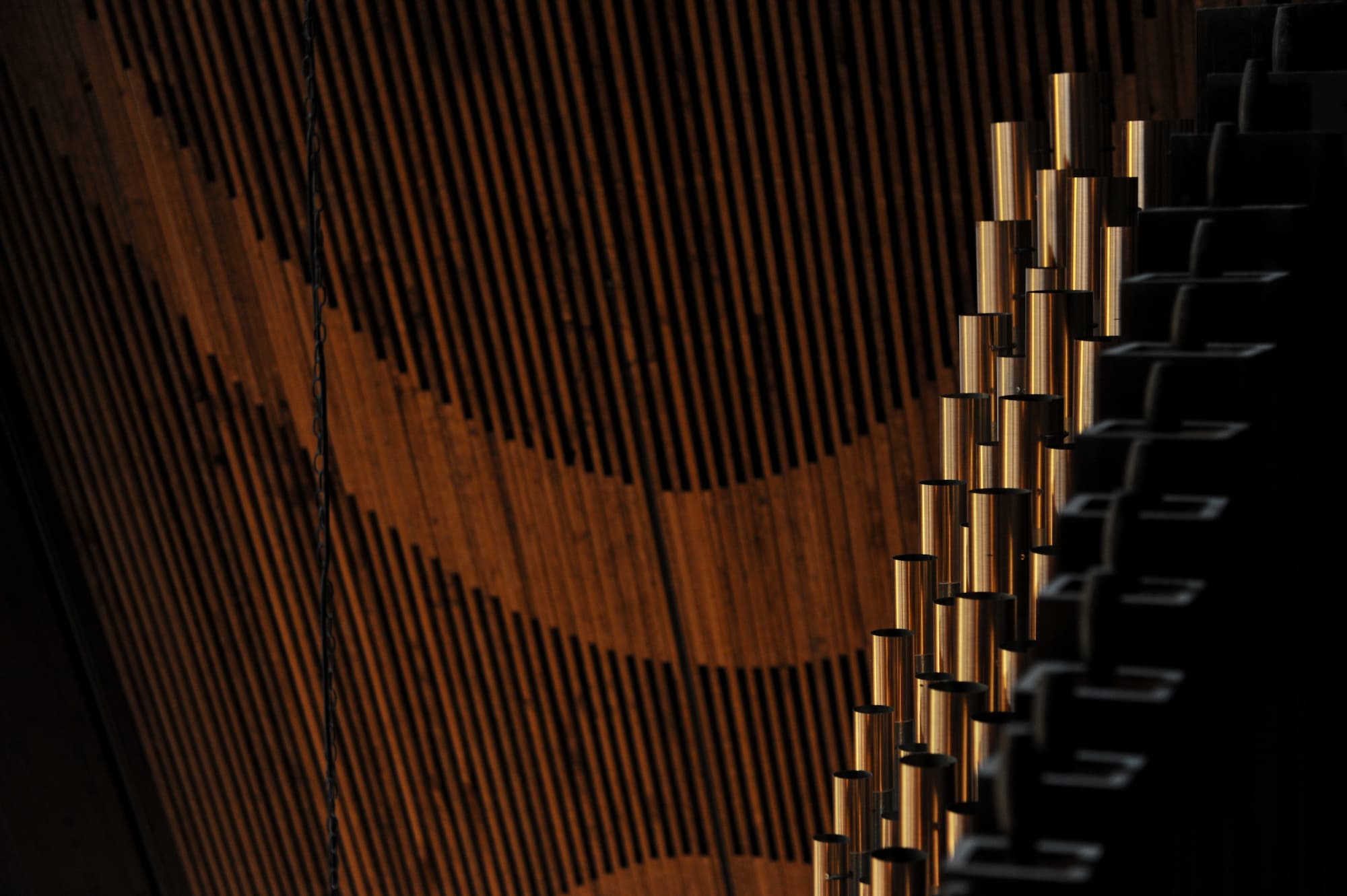 An architectural photograph from the organ loft of Trinity Lutheran Church. In the foreground, warm light catches polished brass pipes from the Wicks 10-rank organ, their sheen contrasting with deep shadows. The background features rhythmic wooden ceiling battens curving along the A-frame roof, blending musical craft with mid-century modern design.