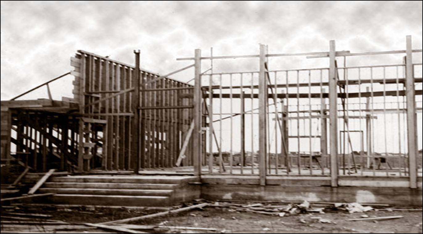 Historic black and white photograph of the wooden structural framing and concrete entrance stairs during the construction of Trinity Lutheran Church's first parish hall in 1955. The image shows the vertical wall studs and skeleton of the multipurpose building against a cloudy sky.