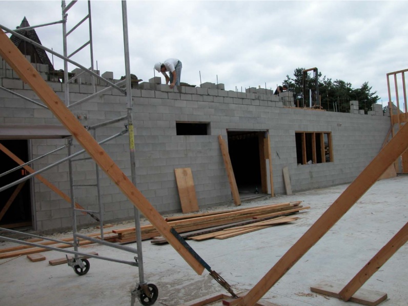 A photograph showing the concrete block masonry construction for the new multi-use facility at Trinity Lutheran Church in Richmond, BC. The southern elevation of the 1964 A-frame church is visible in the background, showing the original roof ridge of the 1964 church building.