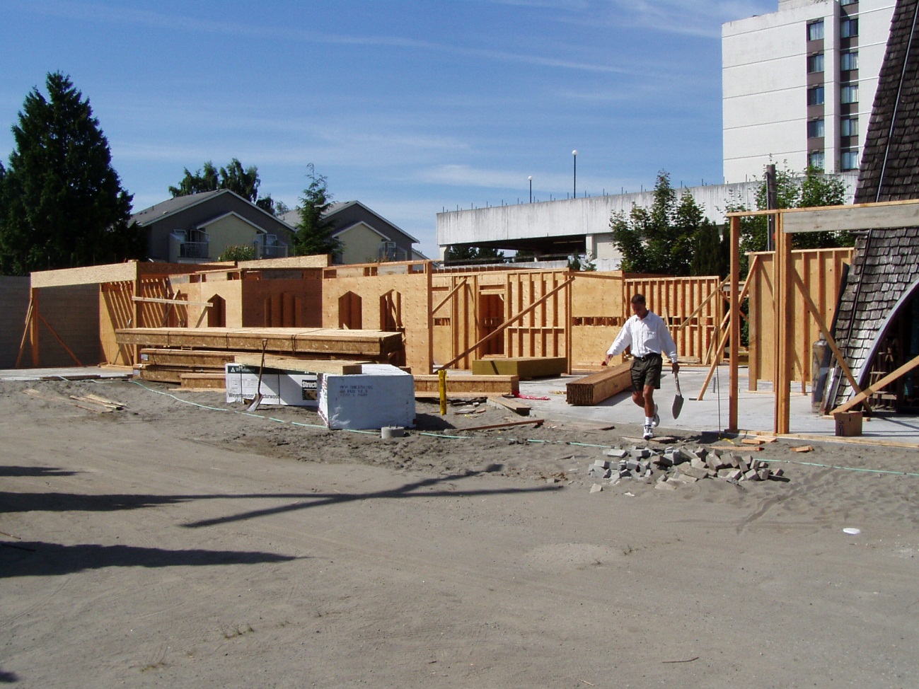 A photograph showing the early stages of building enclosure for the new multi-use facility at Trinity Lutheran Church in Richmond, BC. The image highlights the poured-at-grade concrete slab, the installation of wooden wall framing, and the application of plywood sheathing on the exterior walls.