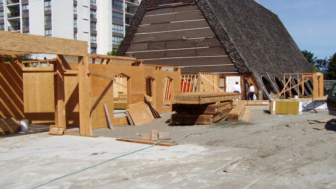 A photograph showing wooden wall framing of the new multi-use facility at Trinity Lutheran Church in Richmond, BC. The existing A-frame church is visible in the background.