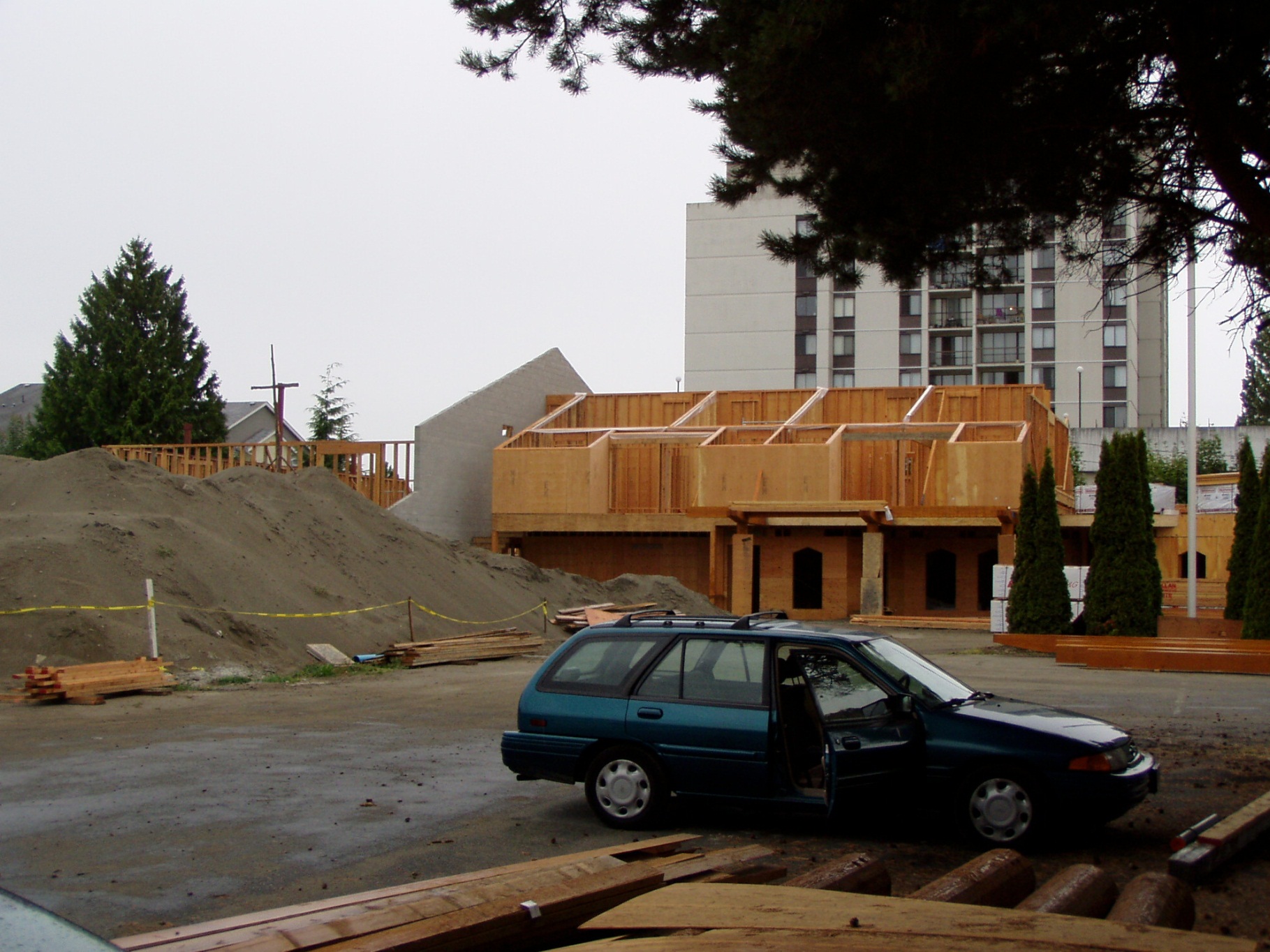 A wide-angle photograph of the construction site for the new multi-use facility at Trinity Lutheran Church in Richmond, BC. The image shows a large pile of preloading sand in the foreground, the concrete block masonry of the parish hall, and the wooden second-floor framing of the main building under a cloudy sky.