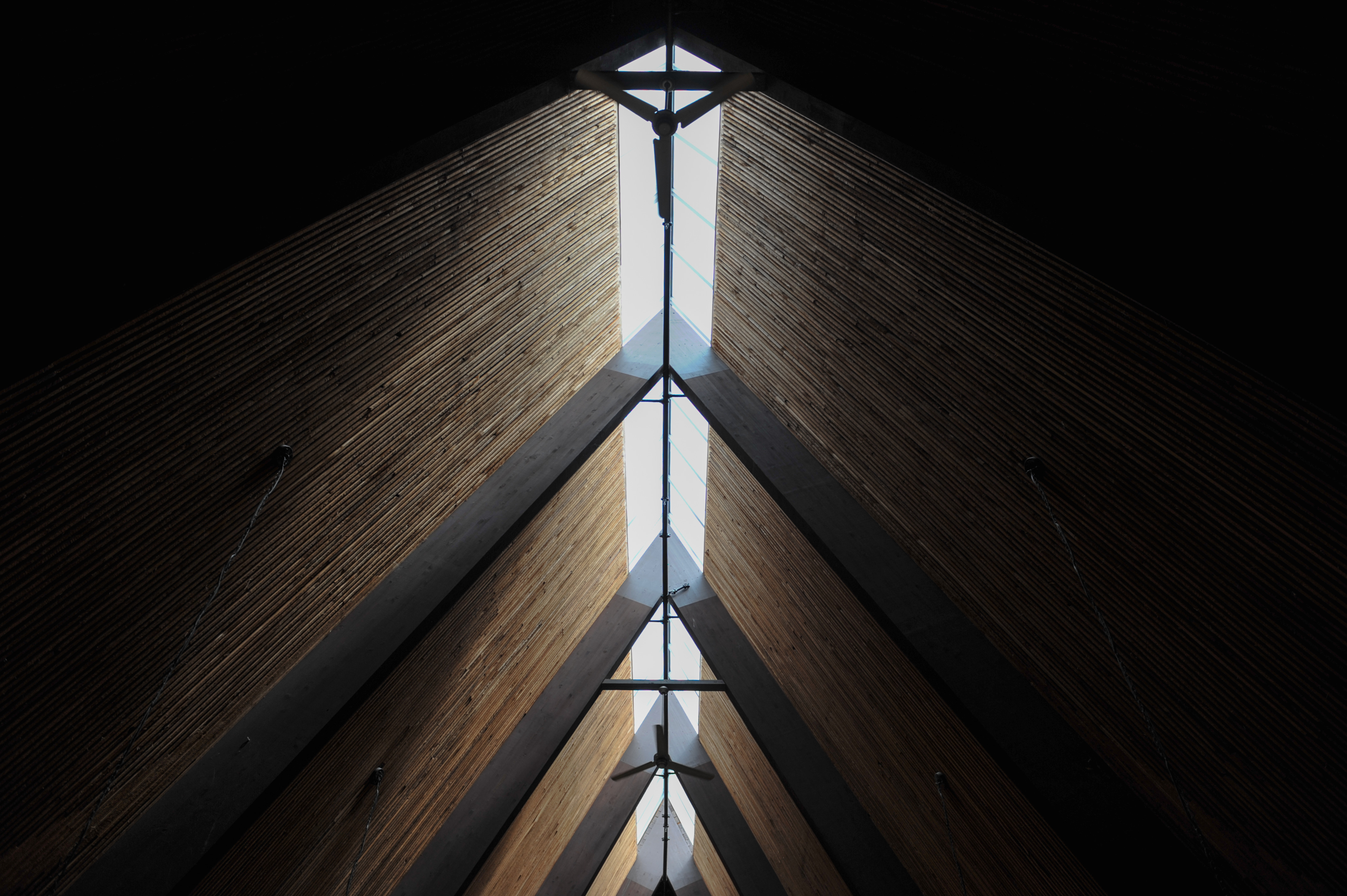 A dramatic architectural perspective looking directly up at Trinity Lutheran Church’s high, vaulted A-frame interior ceiling. A narrow skylight runs the length of the ridge, casting a bright line of natural light that bisects the steeply pitched ceiling. The repeating patterns of dark structural beams and warm wooden slats create a sense of symmetry and soaring vertical space.