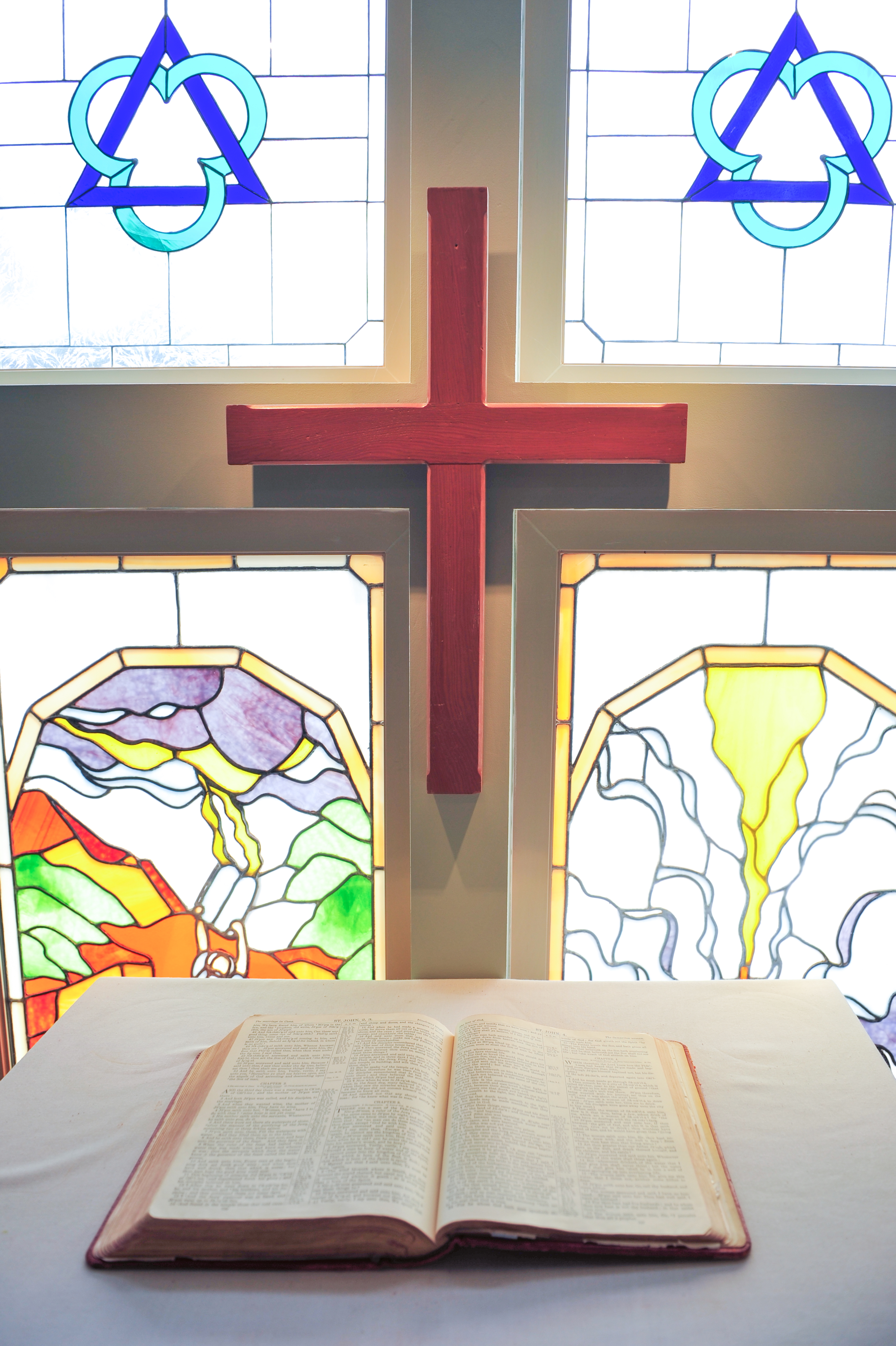 An open Holy Bible resting on a white altar cloth in the chapel. Behind the altar is a wooden cross and stained-glass panels depicting the Holy Trinity, Moses at Mt. Sinai (the Law), and the Crucifixion at Mt. Calvary (the Gospel).