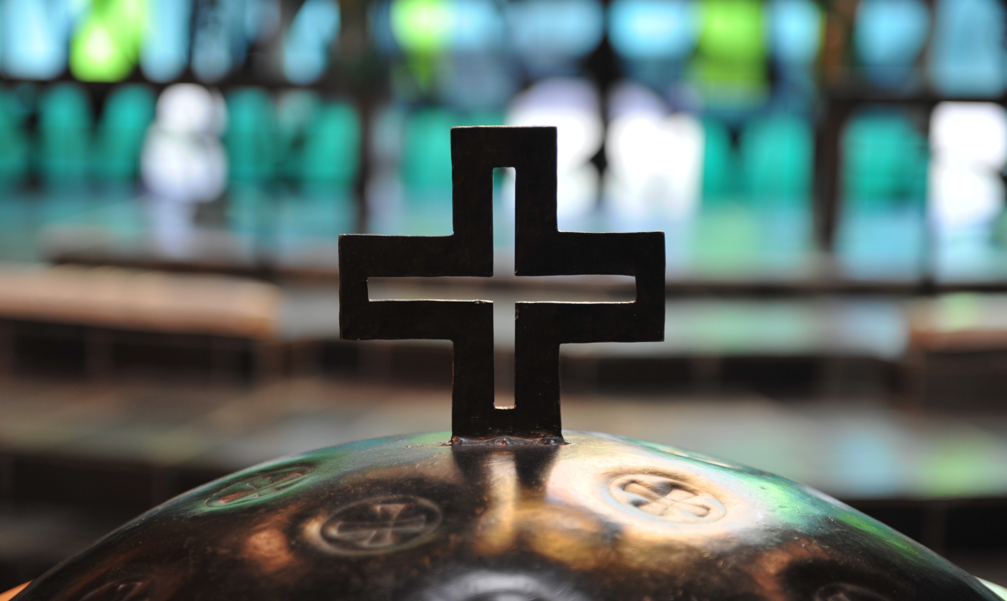 Close-up of the metal baptismal font cover with cross-shaped handle at Trinity Lutheran Church in Richmond, BC. The blurred background shows the chancel and vibrant A-frame stained glass.