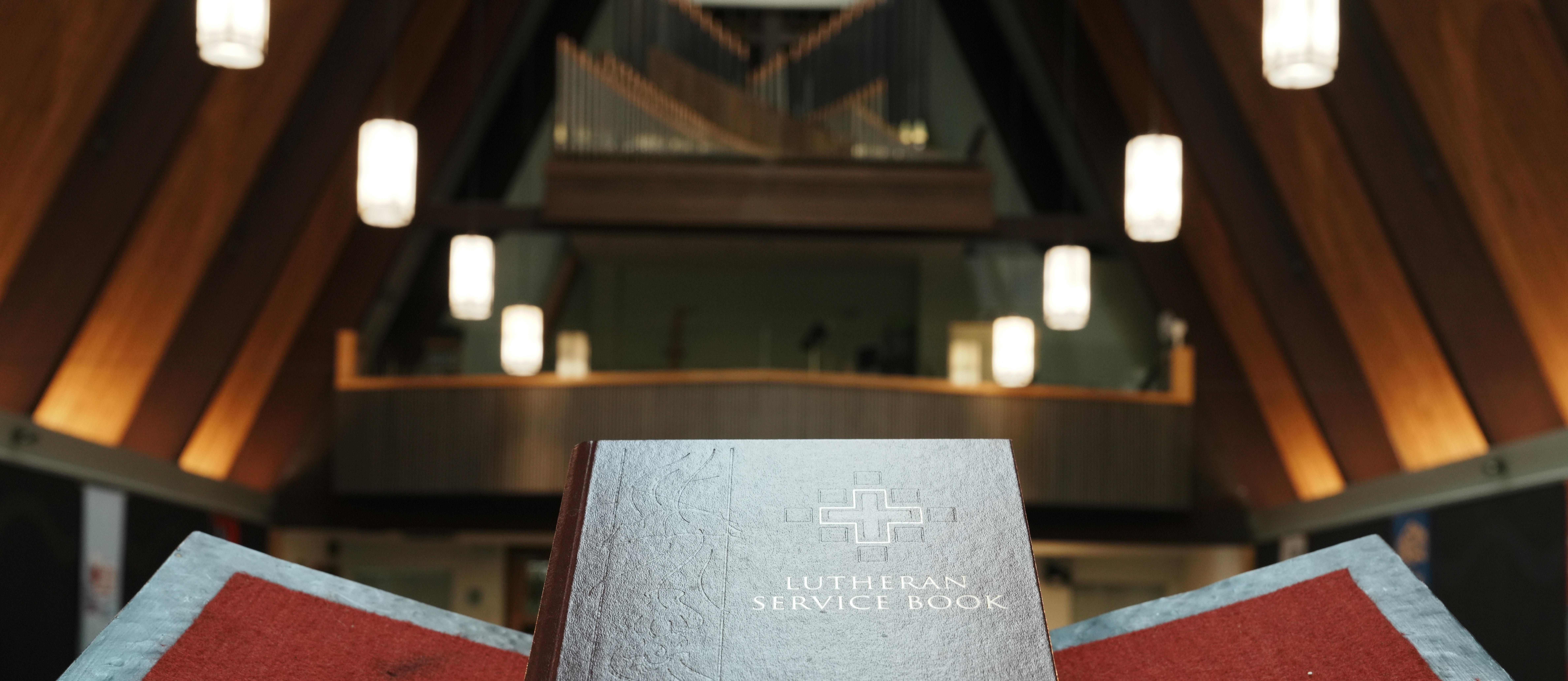 Close-up of the burgundy Lutheran Service Book Altar Book on a stand, containing the historic liturgy for the Divine Service. The background features the warm wooden A-frame nave and pipe organ under Romanesque pendant lights